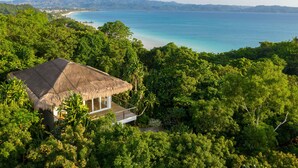 A thatched-roof bungalow with a wooden deck, surrounded by dense tropical foliage, overlooking a clear blue sea and distant hills.