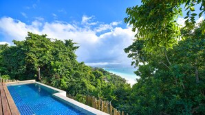 A pool with a wooden deck, surrounded by lush greenery and a view of a beach.