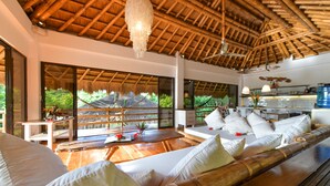 A spacious room with a bamboo ceiling, large windows offering a view of greenery, a wooden sofa with white cushions, and a dining area with a water dispenser.