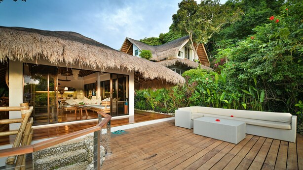 A wooden deck with a thatched-roof structure, white outdoor furniture, and lush greenery.