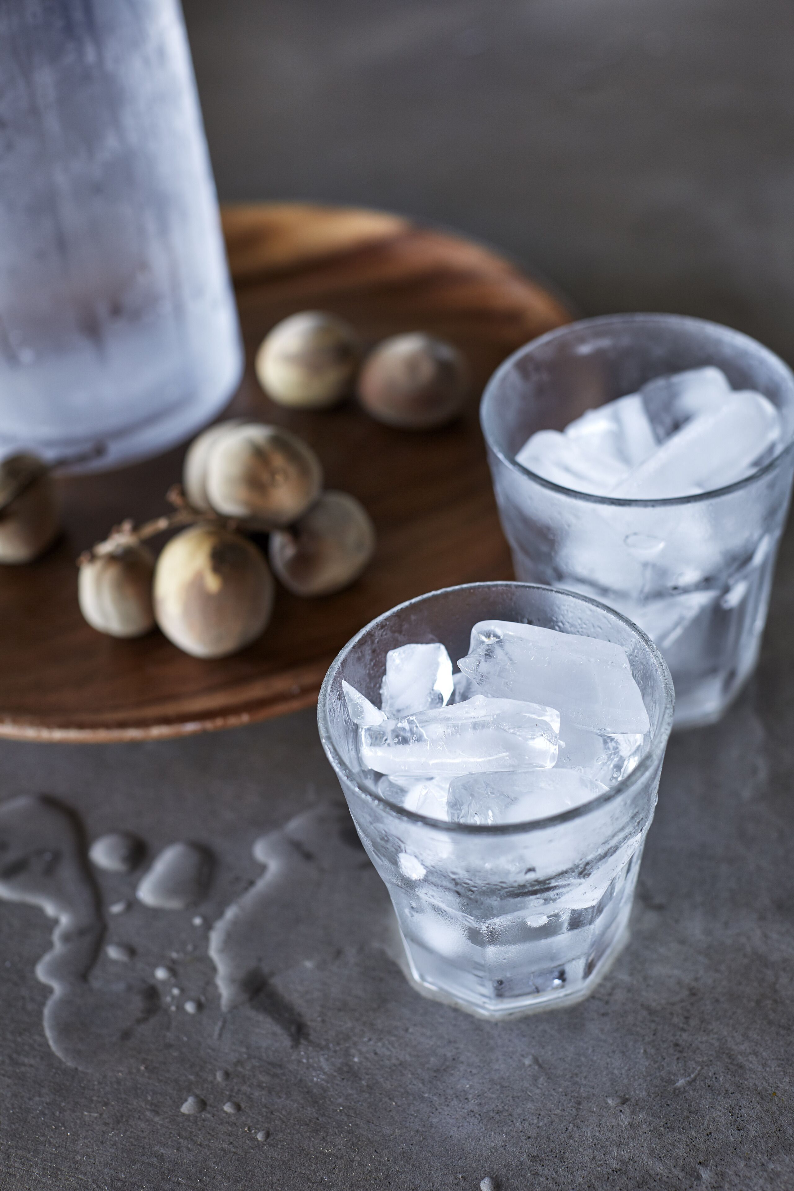 Two glasses of water with ice cubes on a wooden surface, a bottle of water, and some hazelnuts in the background.