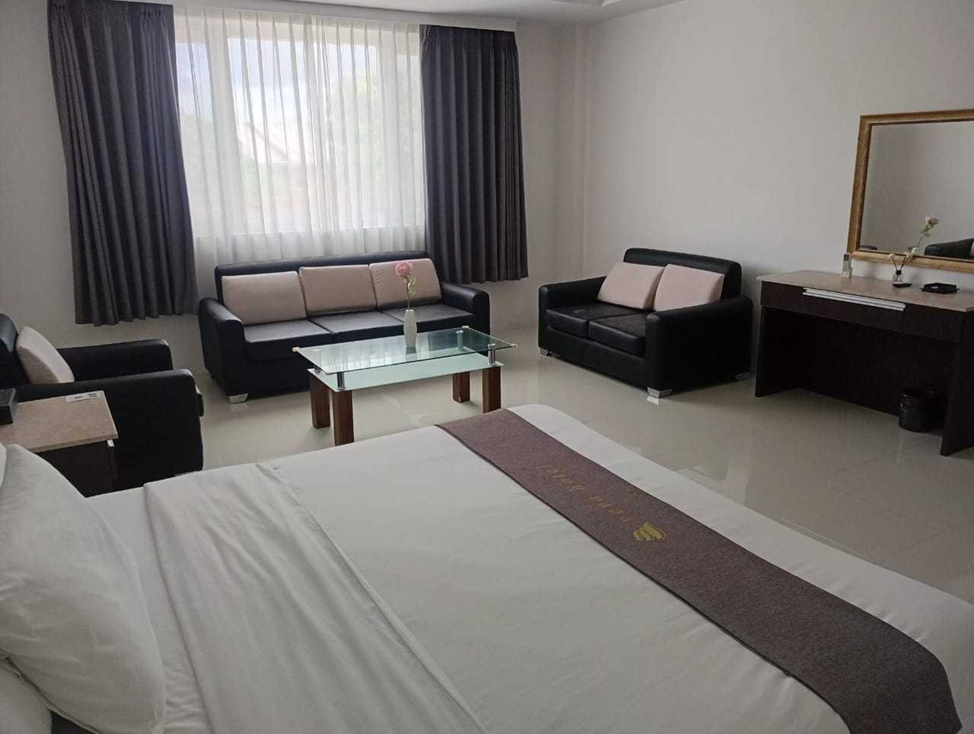 A modern hotel room with a bed, a glass coffee table, two sofas, a desk, and a large window with curtains.