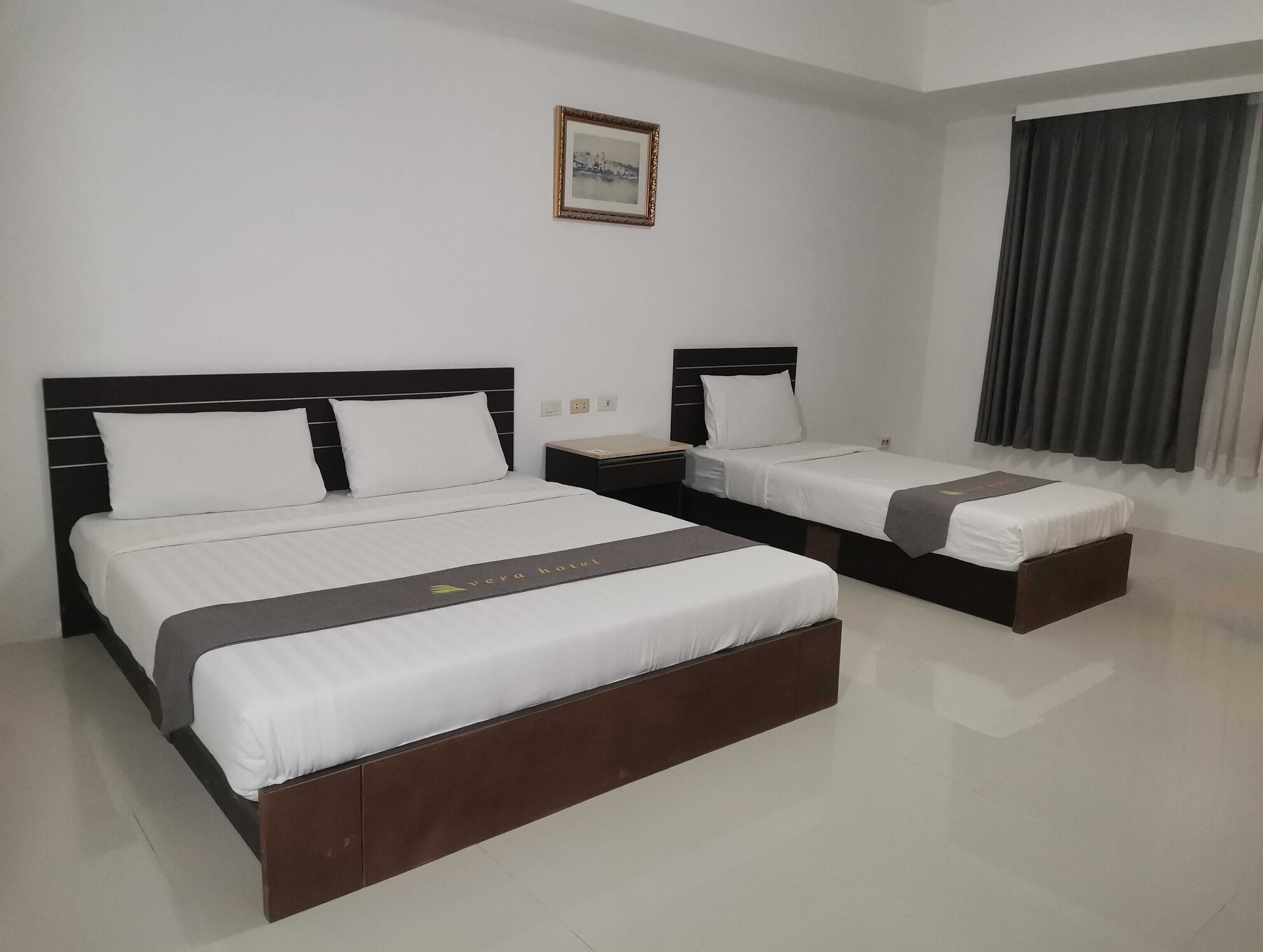Habitación de hotel con dos camas, una mesita de noche, un cuadro enmarcado en la pared y una ventana con cortinas.