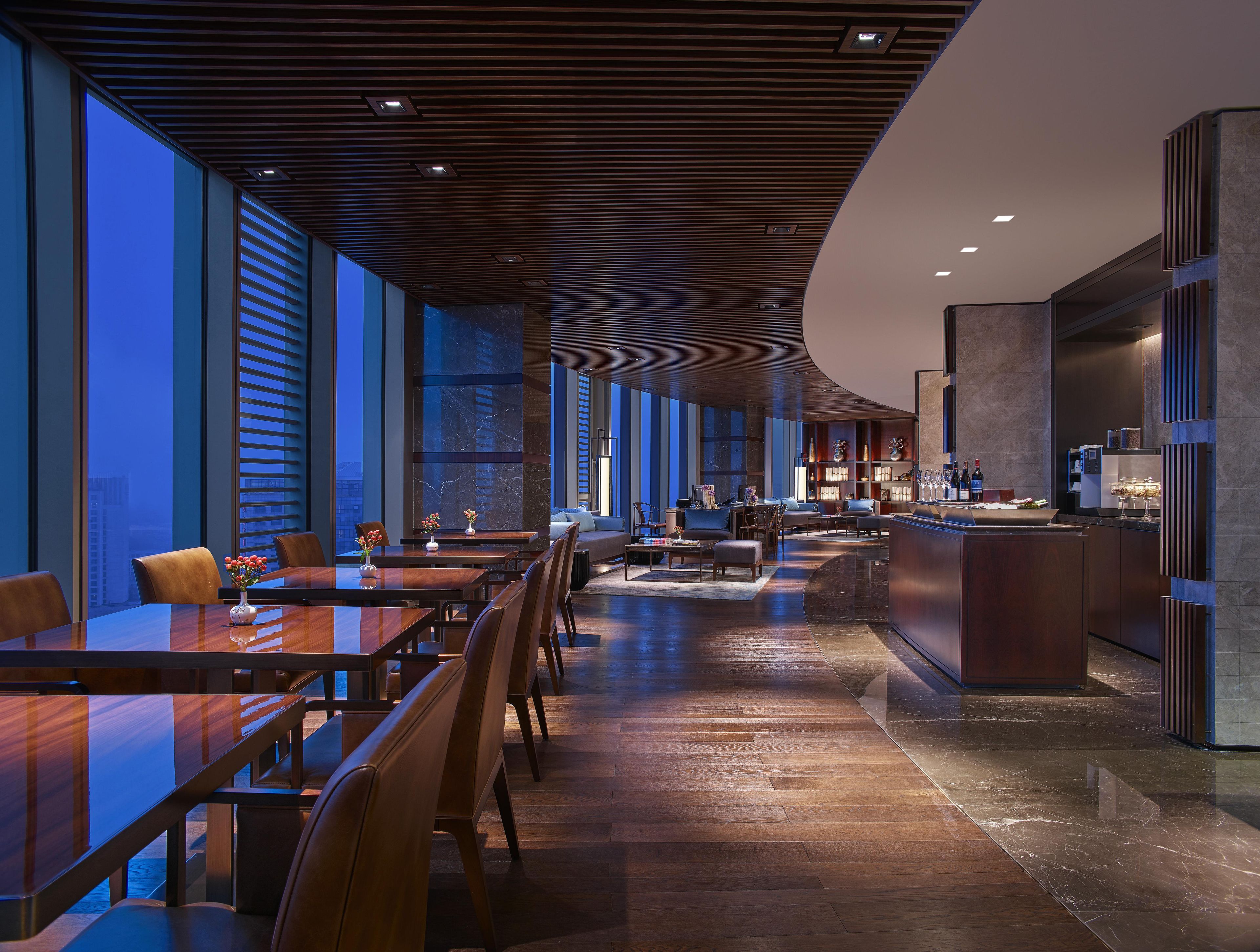 A modern hotel lobby with a long dining area, a bar, and a seating area with a view of the city.