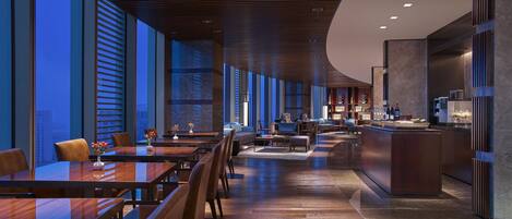 A modern hotel lobby with a long dining area, a bar, and a seating area with a view of the city.