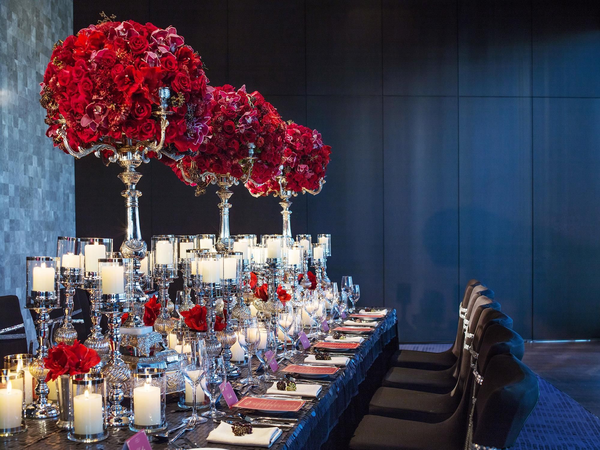 Una larga mesa de comedor dispuesta con múltiples velas, cristalería y flores rojas en jarrones.