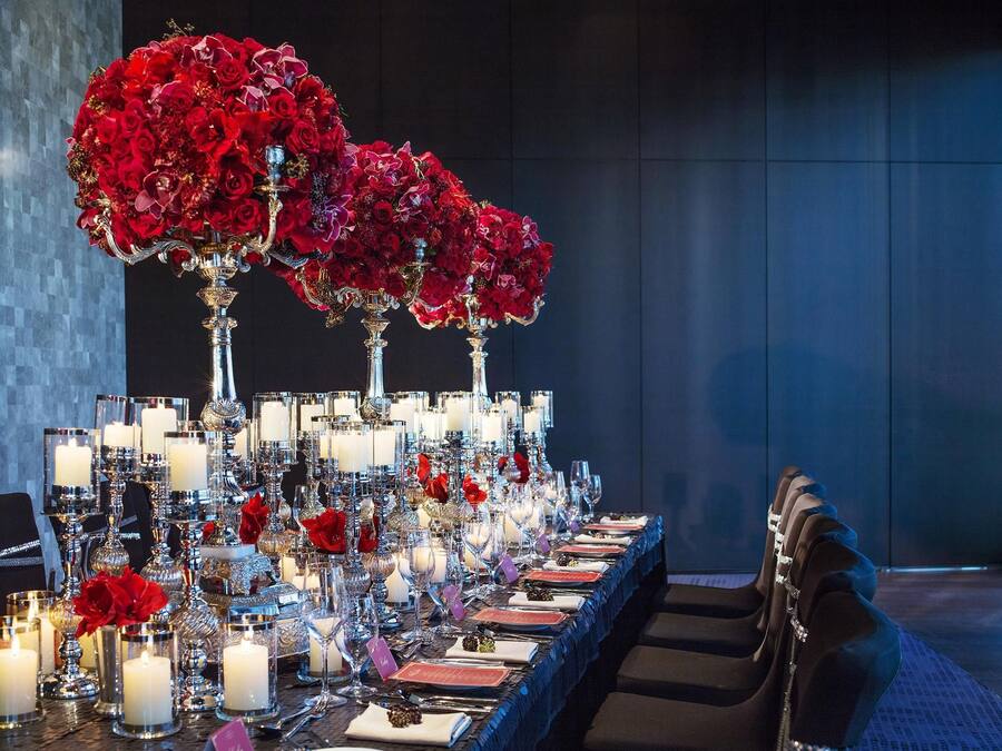 Una larga mesa de comedor dispuesta con múltiples velas, cristalería y flores rojas en jarrones.