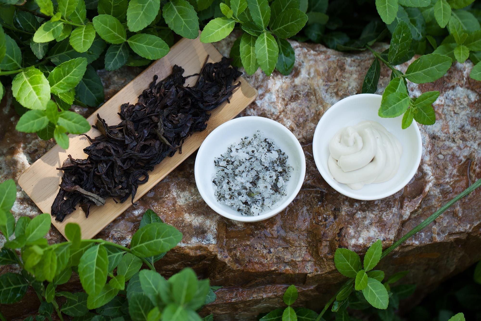 Dried tea leaves, a bowl of salt, and a bowl of cream on a rock surrounded by greenery.