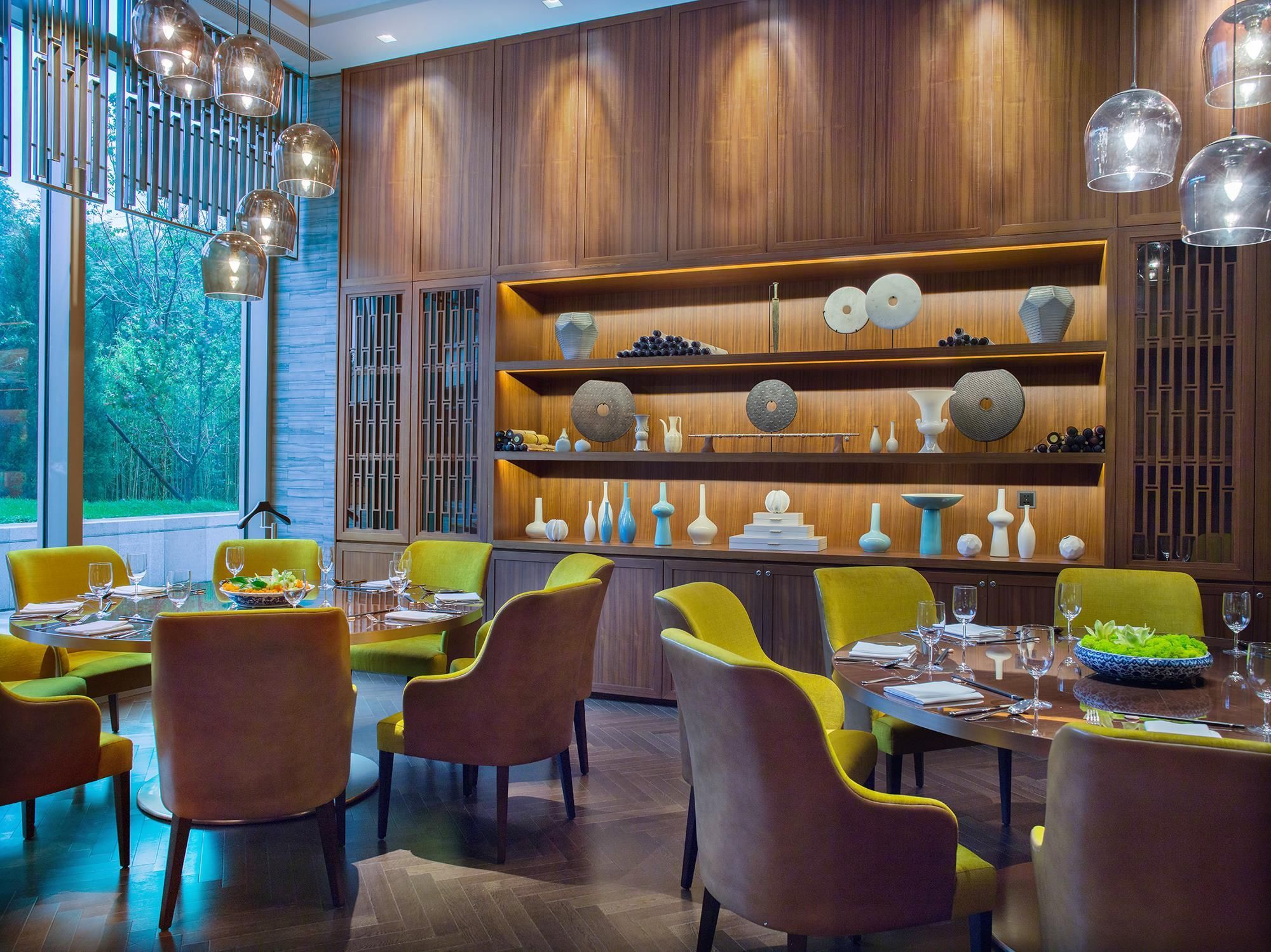 A dining area with yellow chairs, a wooden wall with shelves displaying various decorative items, and a view of trees through large windows.