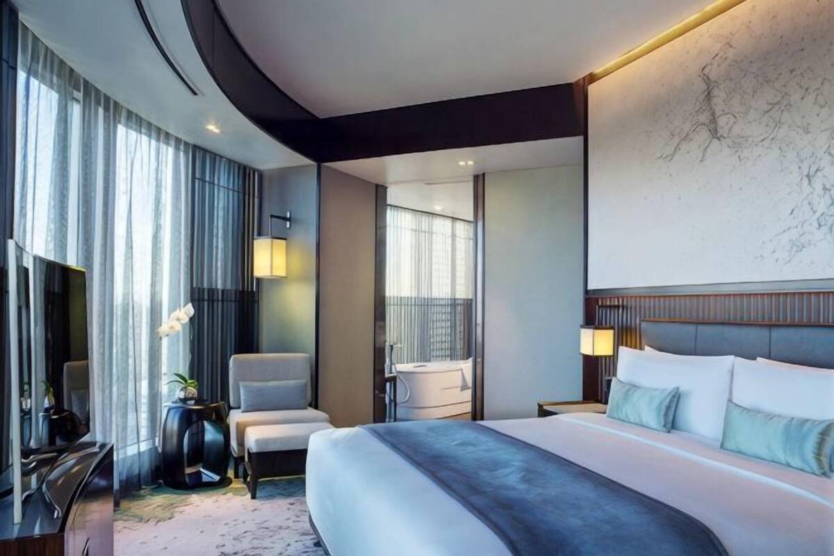 A modern hotel room with a large bed, a sitting area with a chair, a desk, and a large window.