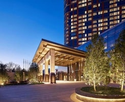A modern hotel entrance with a covered waiting area, a large building in the background, and a landscaped garden.