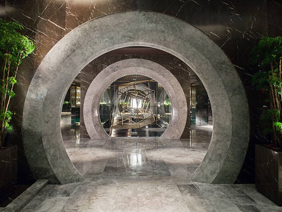 Una estructura arquitectónica circular con un espacio central abierto, rodeada de follaje y superficies reflectantes.