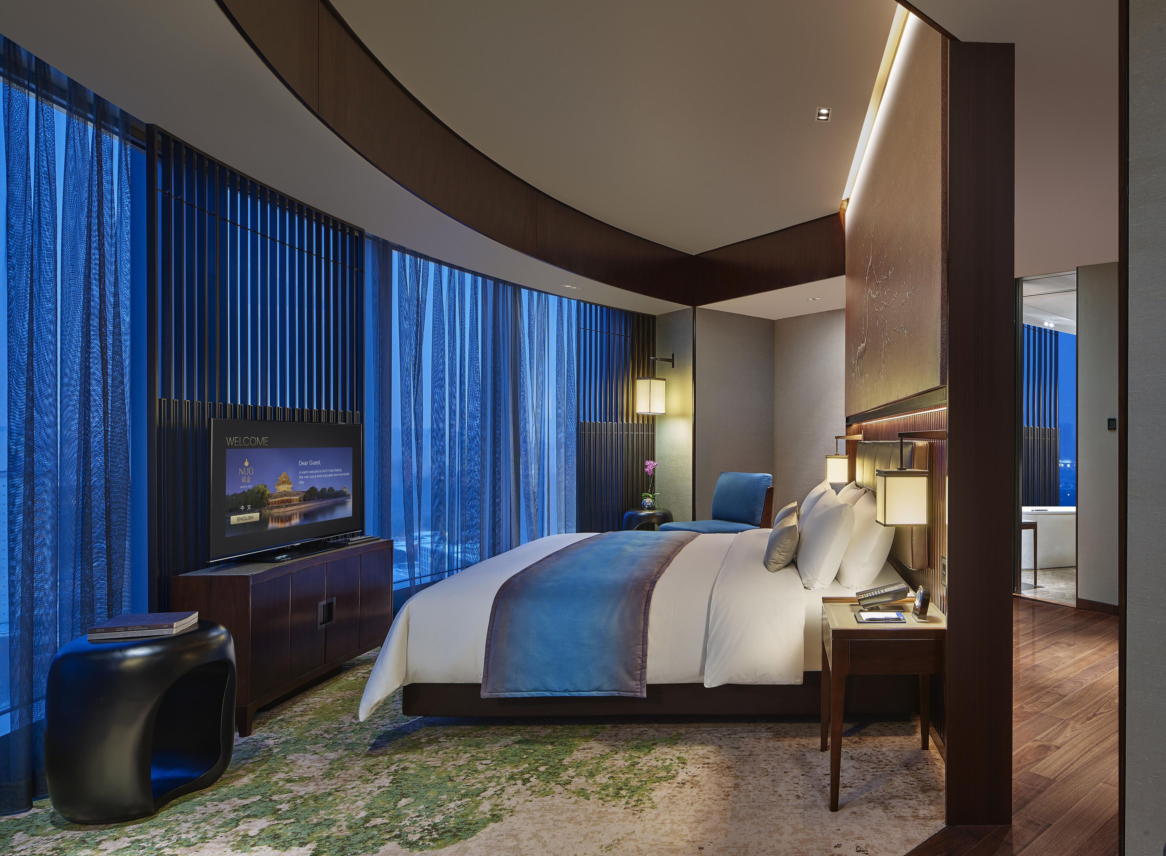 A modern hotel room with a large bed, a television, and a view of the cityscape.
