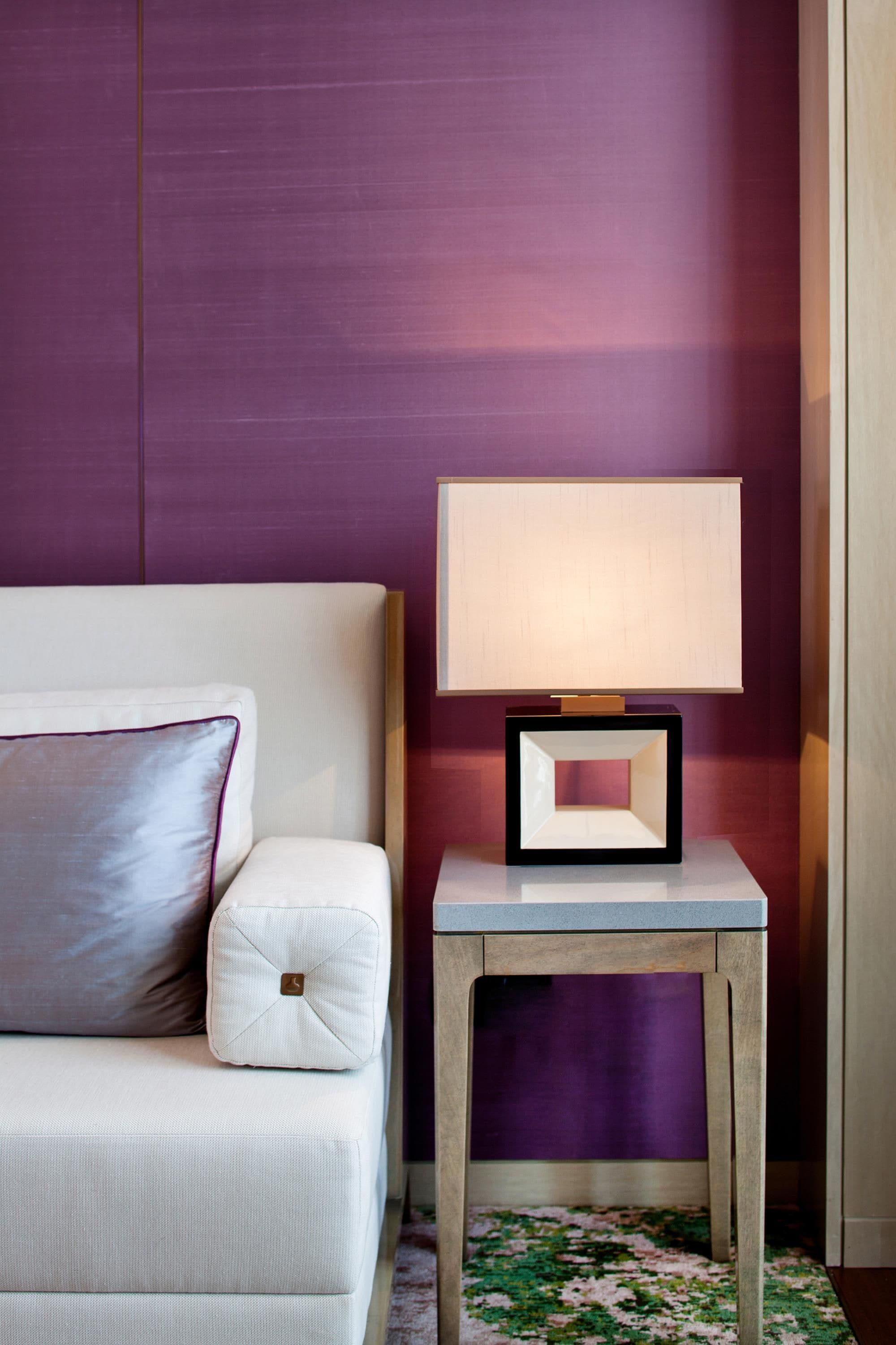 A hotel room with a white sofa, a bedside table with a lamp, and a purple wall.