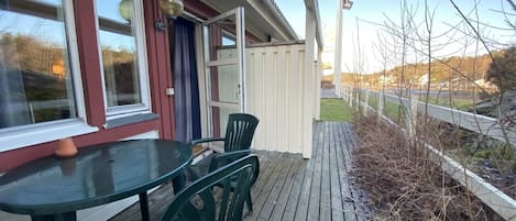 En træterrasse med et bord og stole, en dør samt udsigt til et indhegnet område med træer.