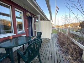 Una terraza de madera con una mesa y sillas, una puerta y vistas a un área cercada con árboles.