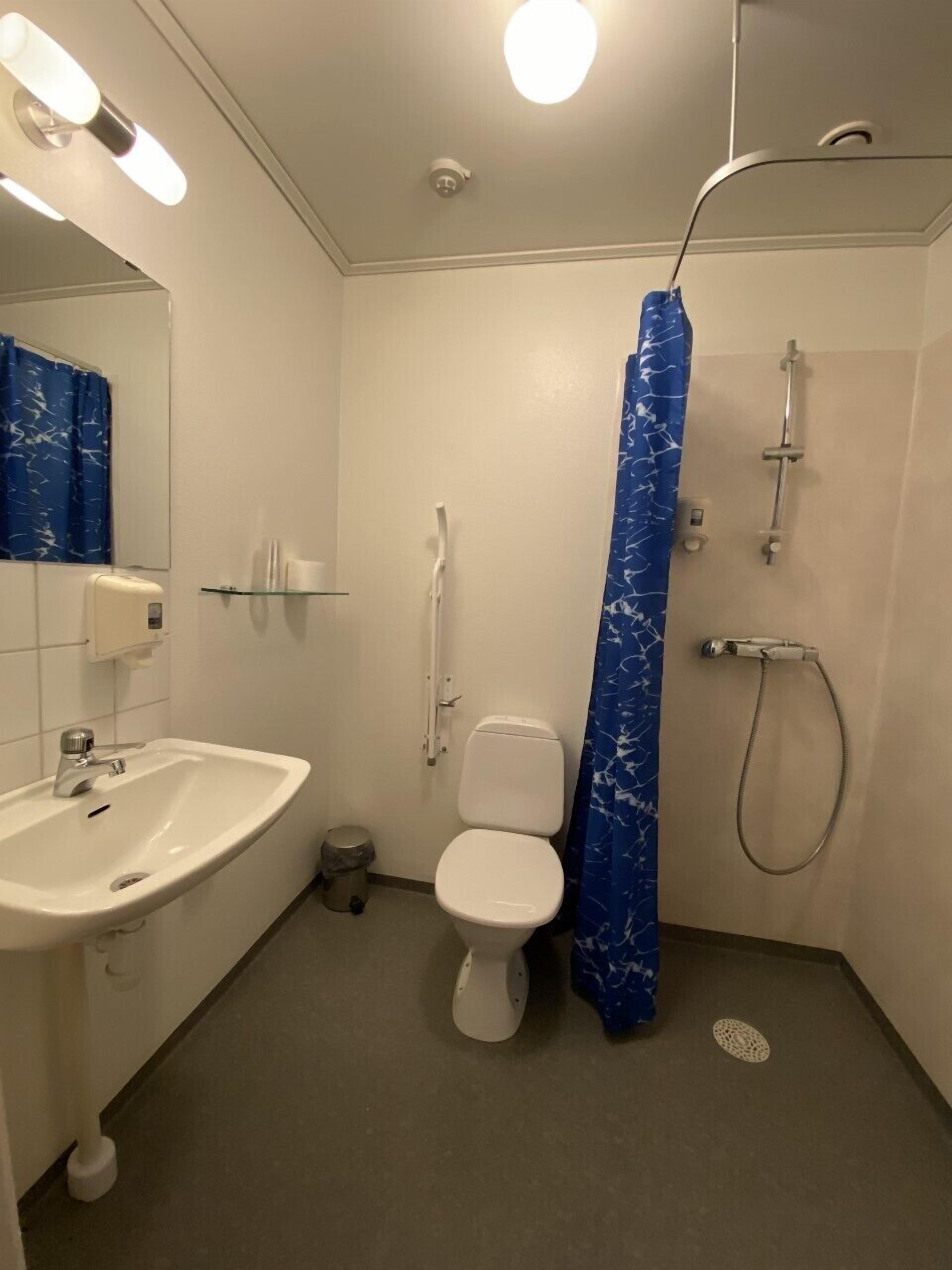 Egy fürdőszoba, amelyben fehér mosdókagyló, WC és zuhanyzó található. Kék színű zuhanyfüggöny és egy üvegpolc van a mosdókagyló felett.