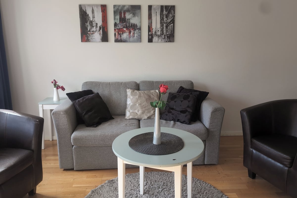 Un salón con un sofá gris, dos sillones negros, una mesita blanca con un jarrón de flores y tres cuadros en la pared.
