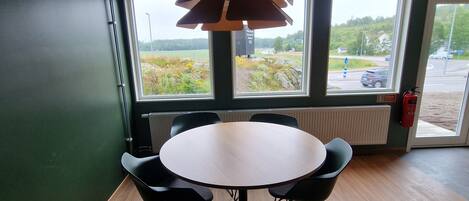 A conference room with a round table and six chairs, large windows with a view of a road and greenery, and a ceiling with a light fixture.
