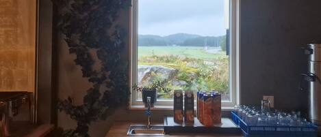 Une cuisine avec une fenêtre donnant sur un paysage rural, un évier et divers récipients sur le plan de travail.