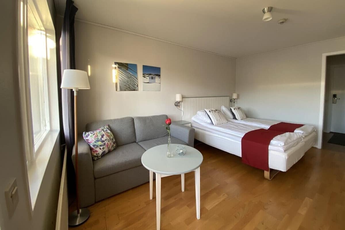 Una habitación compacta con un sofá gris, una cama individual con una colcha roja, una mesita redonda con un jarrón y dos lámparas fijadas a la pared.