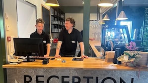 A reception desk with a marble top and the word "RECEPTION" in bold letters. There are two individuals behind the desk, one operating a computer. - Nordby Hotell (Stromstad)
