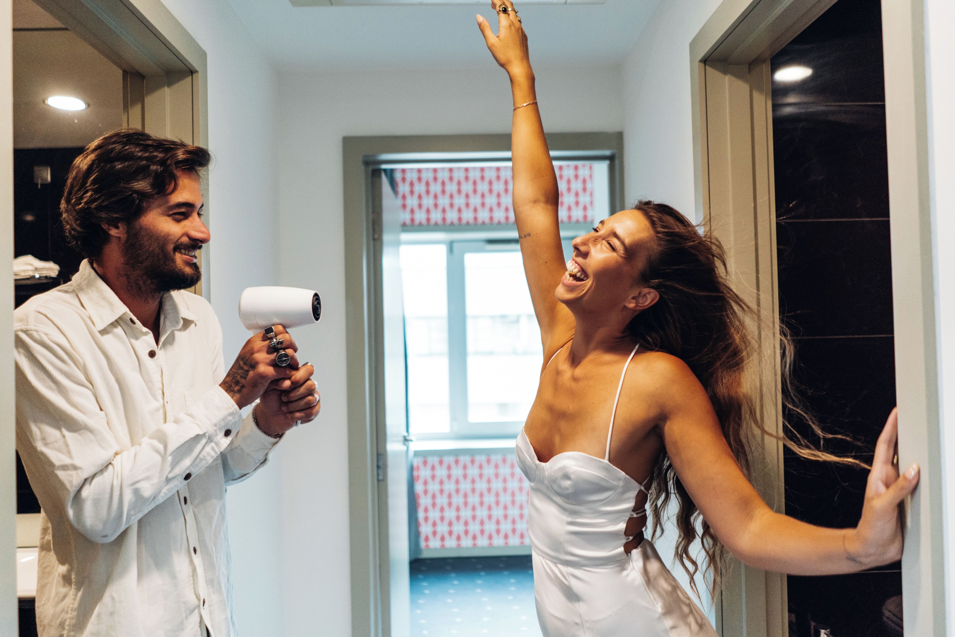 A man holding a hairdryer and a woman with her hair being blow-dried in a hallway with patterned wallpaper and a door in the background.