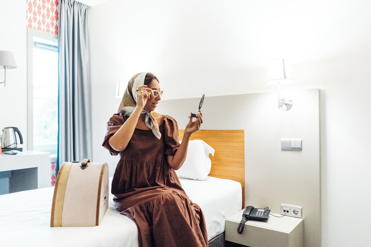 A hotel room with a bed, a bedside table with a lamp, a telephone, and a window with curtains.