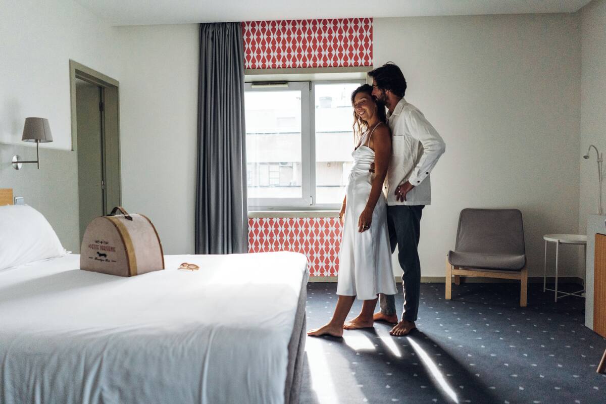 A hotel room with a bed, a suitcase, a chair, and a window with curtains.