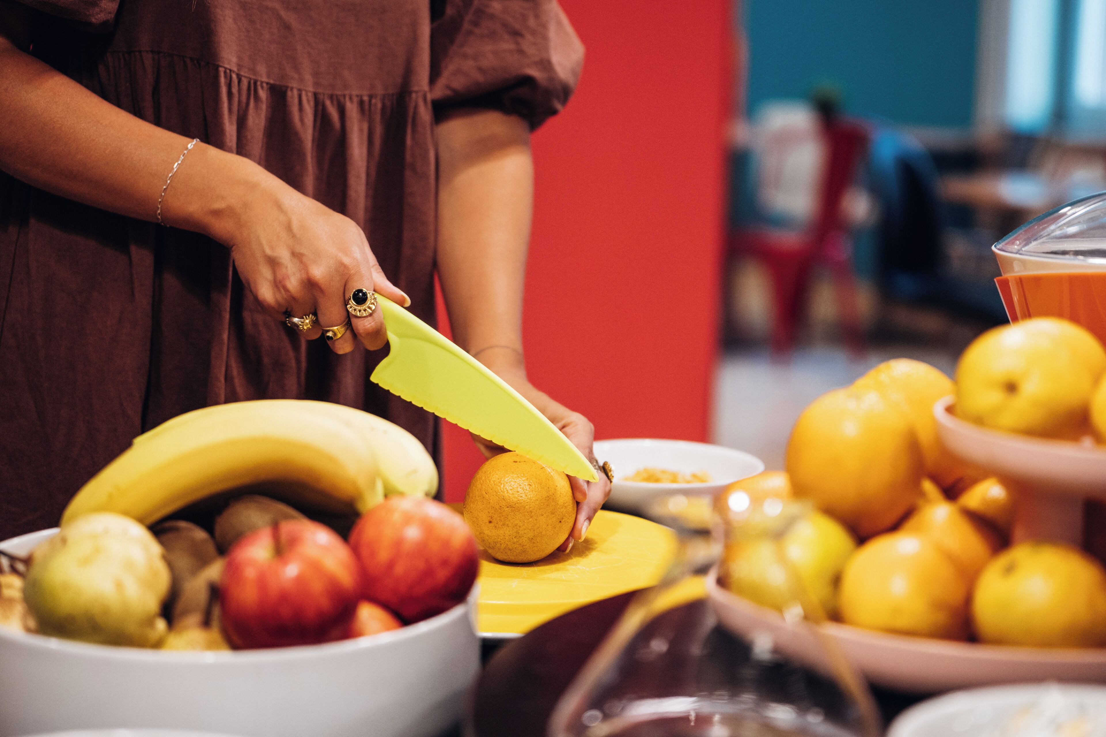 Una persona taglia un'arancia con un coltello giallo su un tavolo pieno di frutta e ciotole.