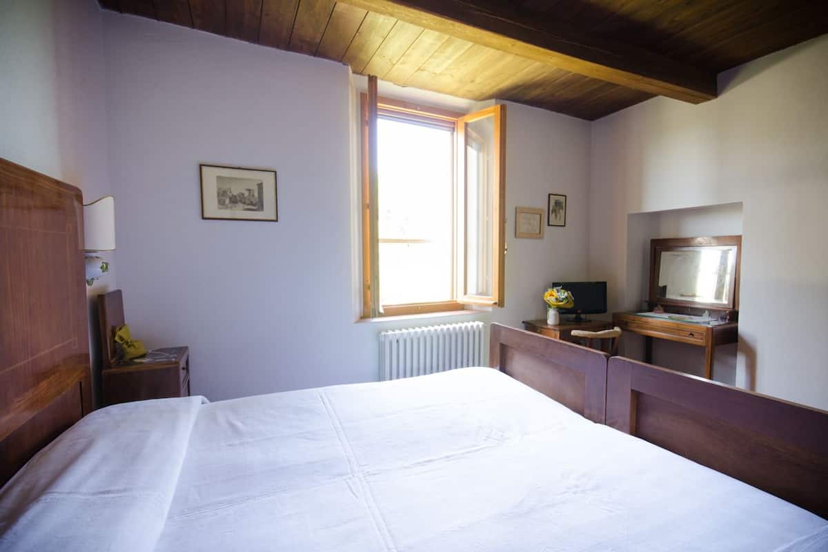 A bedroom with a large bed, a wooden headboard, a desk with a computer, a window with a view, and a radiator.