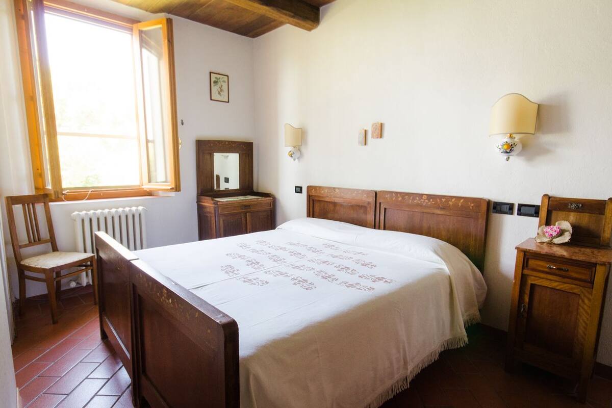 A bedroom with a wooden bed, a chair, a nightstand, a mirror, and a window with a view of trees.