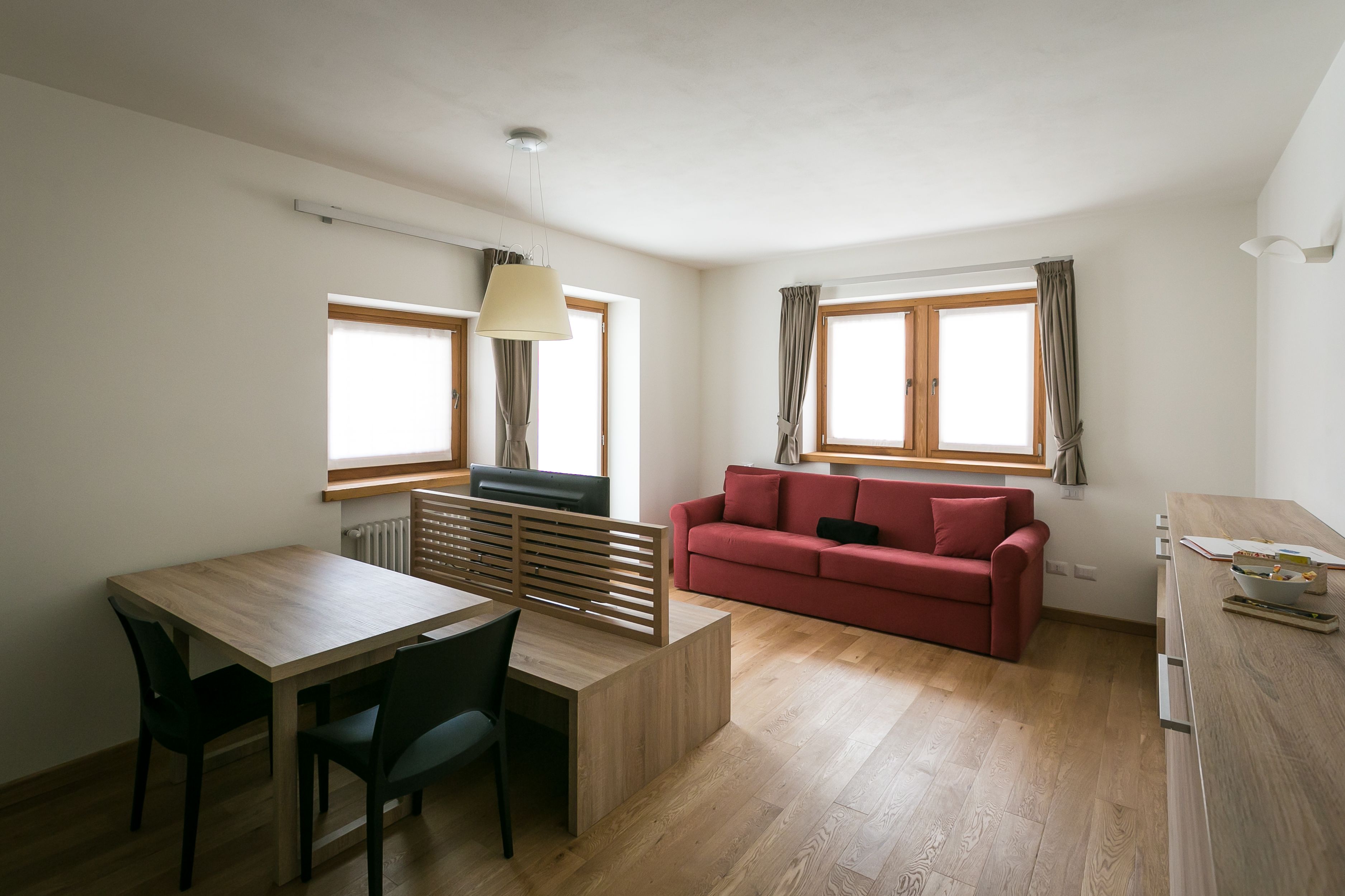 A modern living room with a wooden dining table, a red sofa, and a kitchen area with a wooden countertop.