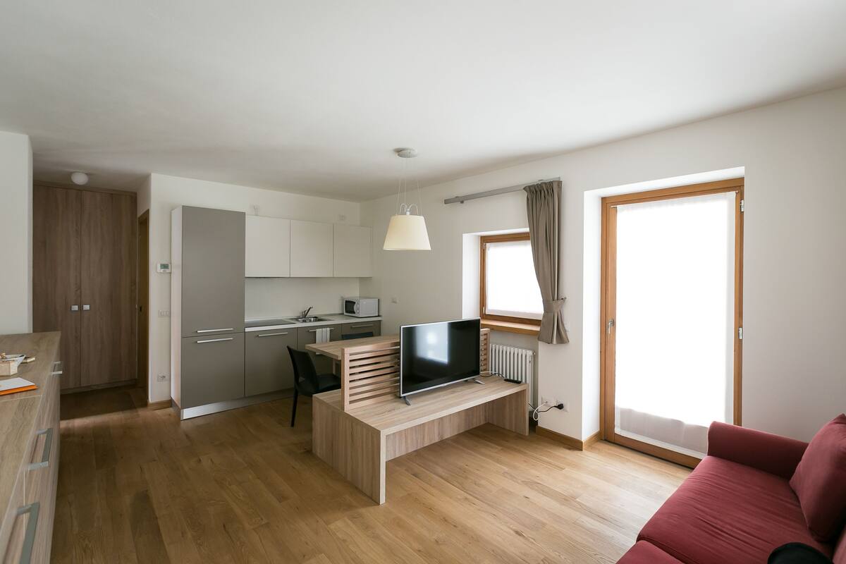 En moderne lejlighed med et køkken, spiseplads, fjernsyn på et træskrivebord og en rød sofa.