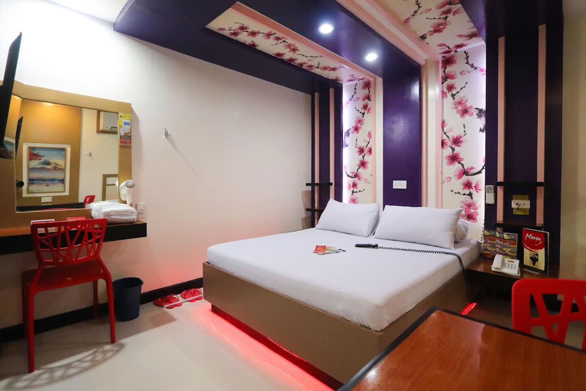 A hotel room with a bed, a desk with a chair, a mirror, and a red chair.