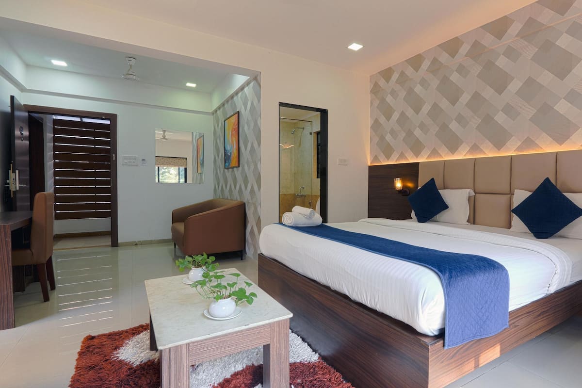 A modern hotel room with a large bed, a desk, and a chair. There is a bathroom visible through a glass door.