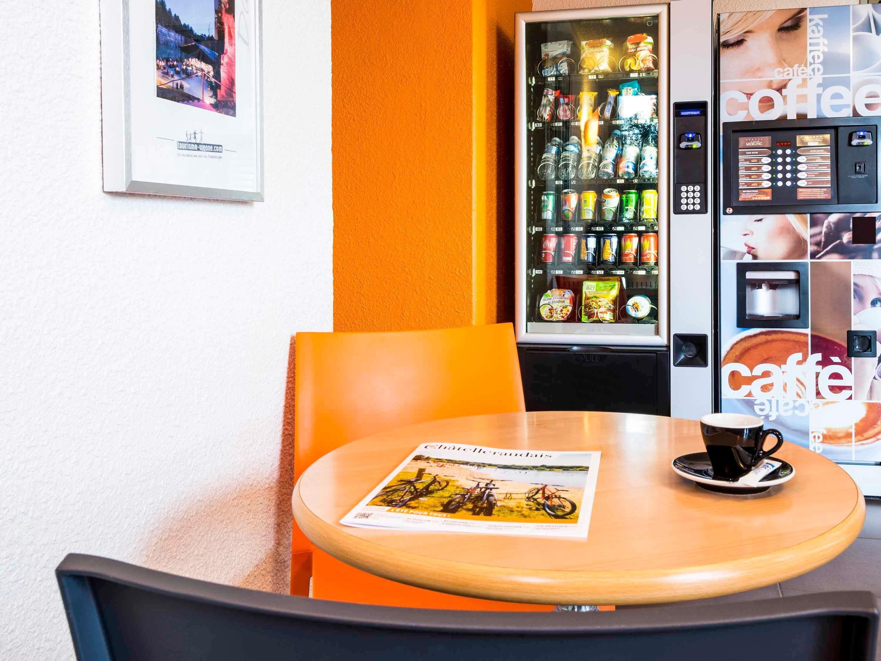 A café with a wooden table, a framed picture on the wall, a vending machine with beverages, and a coffee machine.