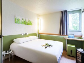 A hotel room with a bed, a small table with a laptop, a green stool, and a window with curtains.