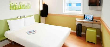 A hotel room with a bed, a desk with a laptop, a TV, a window, and a wall-mounted artwork.