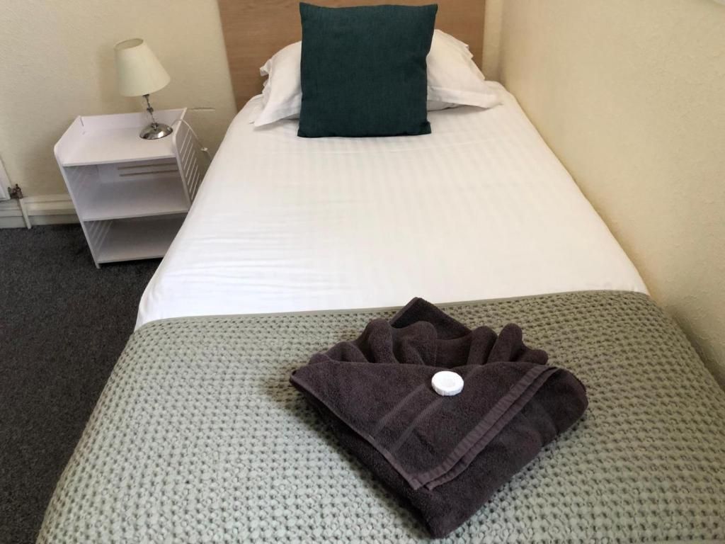 Una cama bien hecha con un almohadón blanco y uno verde decorativo, una toalla oscura doblada sobre una manta gris de punto, y una mesita de noche blanca con una lámpara.