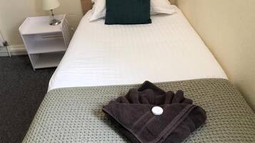 Una cama bien hecha con un almohadón blanco y uno verde decorativo, una toalla oscura doblada sobre una manta gris de punto, y una mesita de noche blanca con una lámpara.
