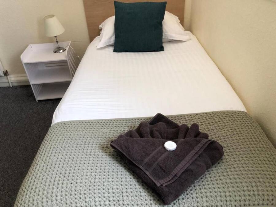 Una cama bien hecha con un almohadón blanco y uno verde decorativo, una toalla oscura doblada sobre una manta gris de punto, y una mesita de noche blanca con una lámpara.