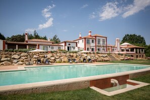 A large swimming pool with a stone border, surrounded by a landscaped garden and a multi-story building with a tiled roof.