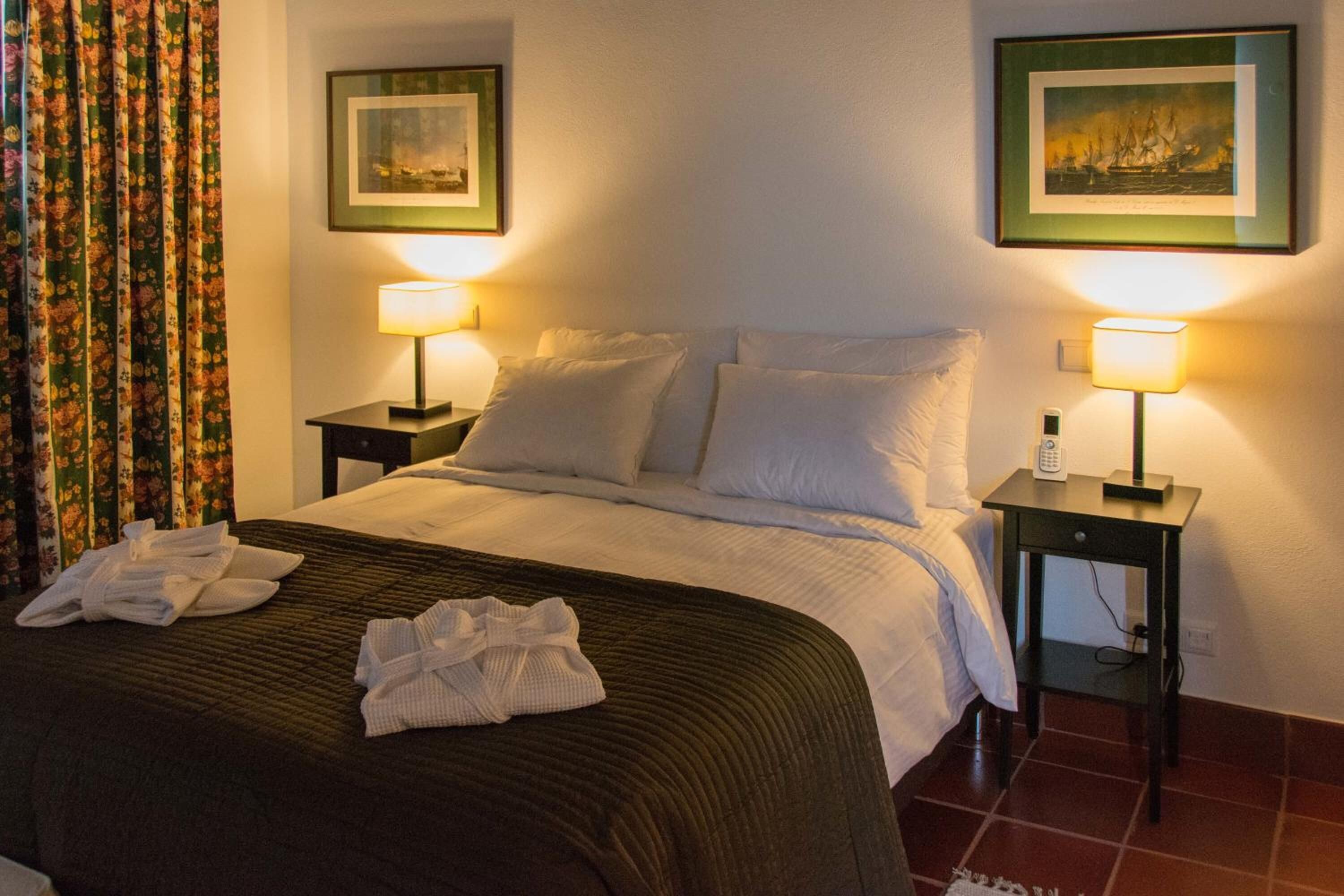 Una cama bien hecha con sábanas blancas, dos mesitas de noche con lámparas y una cortina estampada con flores.