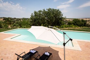 Piscina con dos sillas reclinables y una toldilla, rodeada de vegetación y colinas.