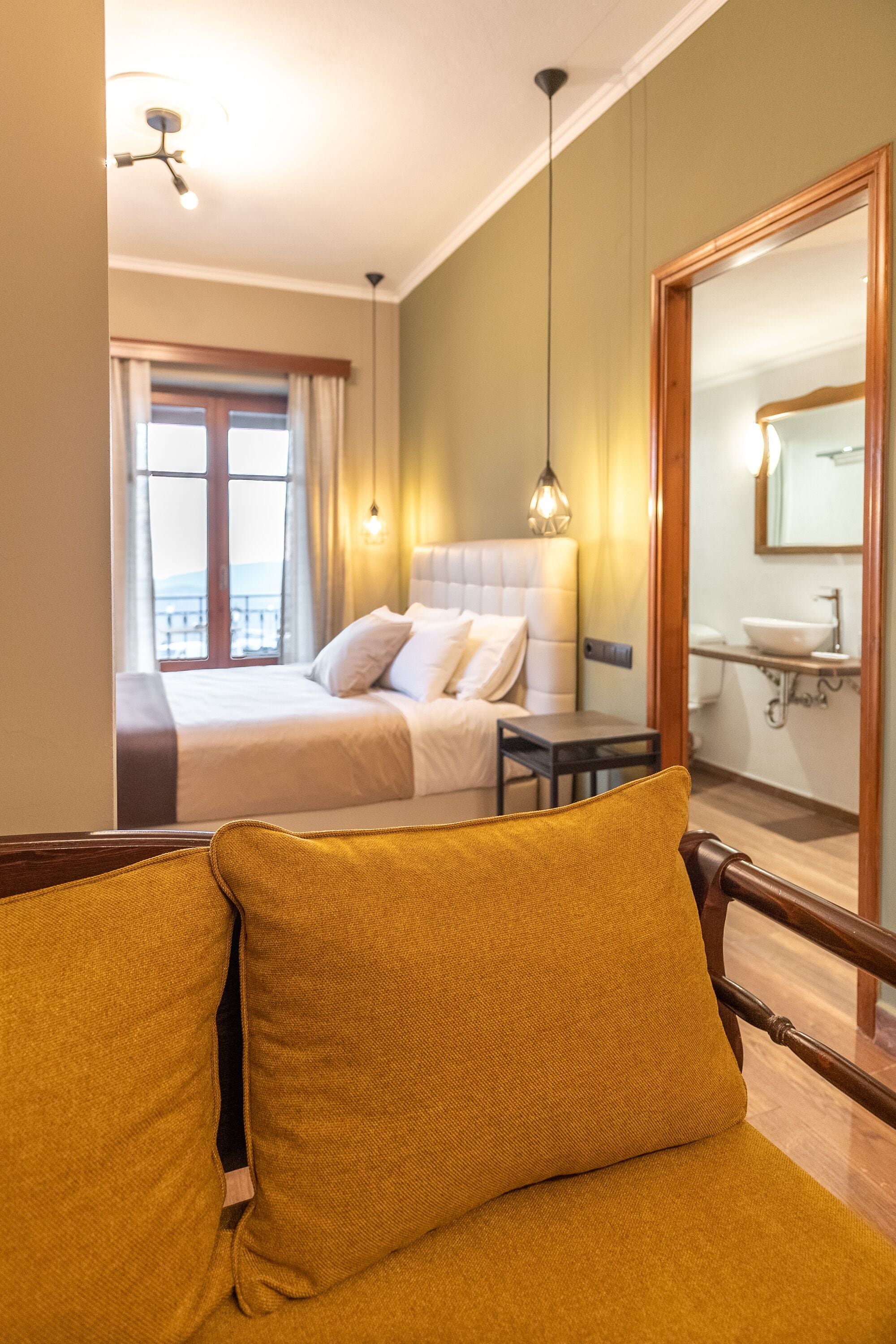 Habitación de hotel con cama, silla, escritorio, espejo y baño visible a través de una puerta abierta.