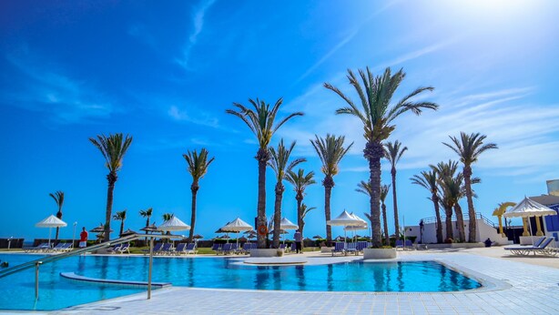 A swimming pool with palm trees and lounge chairs, surrounded by umbrellas and a clear blue sky. - Aljazira Beach & Spa  -  Families and Couples Only (Mezraia)