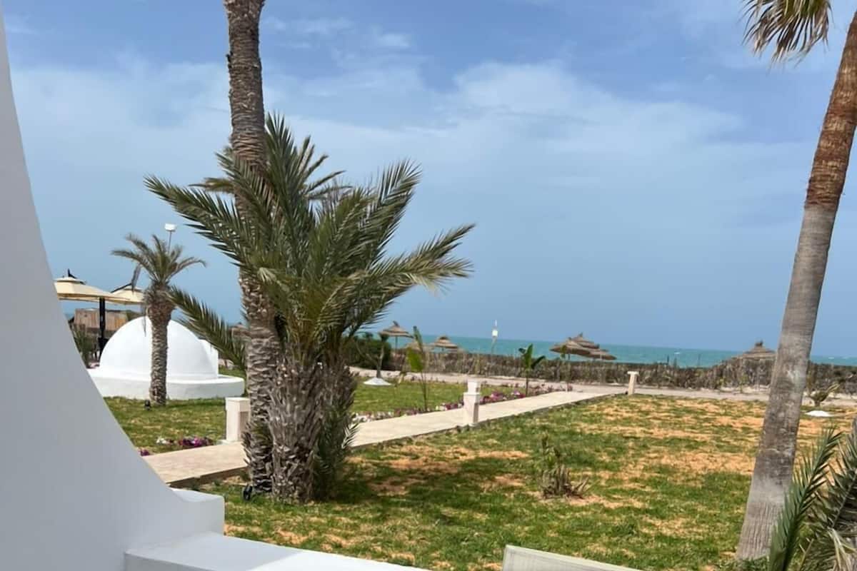 Eine Terrasse mit Tisch und Stühlen, mit Blick auf einen Strand mit Palmen und einer weißen Kuppelstruktur.