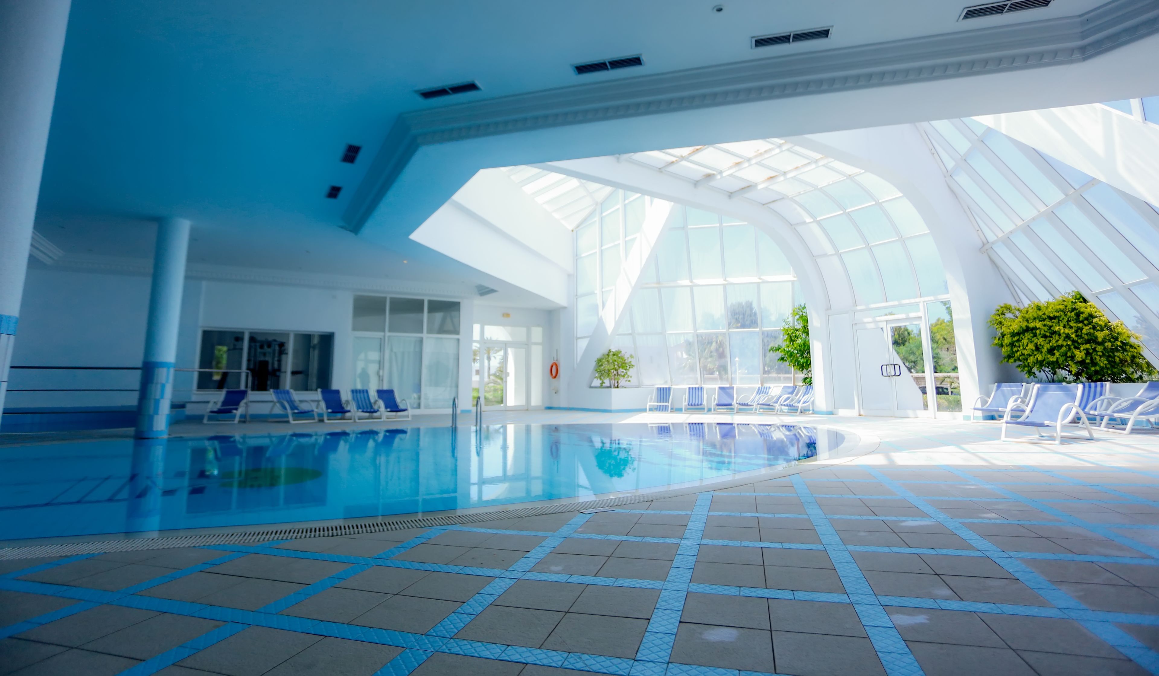 An indoor swimming pool with clear blue water, surrounded by lounge chairs and a glass ceiling allowing natural light.
