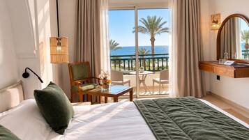 Una camera d'albergo con un letto, una scrivania con una sedia, un tavolino con un vaso di fiori, un balcone con vista sul mare e una lampada a sospensione.